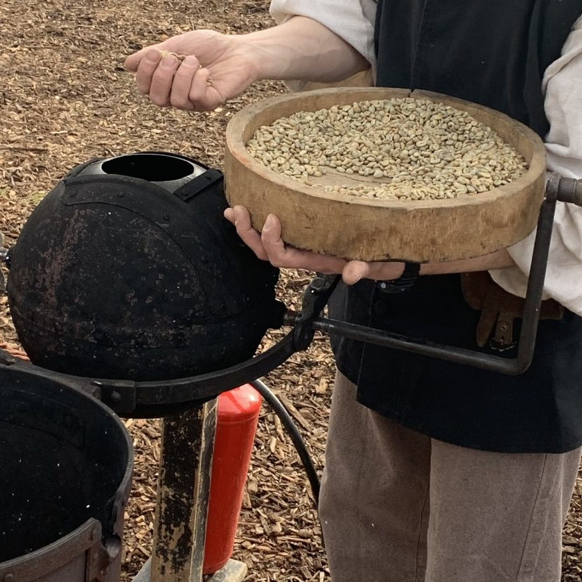Eine Person hält einen hölzernen Sieb mit ungerösteten Kaffeebohnen über einen schwarzen Ofen.